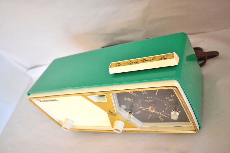 May include: A retro green and cream alarm clock with a gold-coloured trim. The clock face is black with gold hands and numerals. The top of the clock has a white panel with the words "Sleep Saver" and "Dose Alarm".