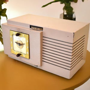 以下が含まれることがあります： ピンクとゴールドのヴィンテージラジオ。時計の文字盤とスピーカーグリルが付いています。ラジオは茶色のテーブルの上に置かれています。