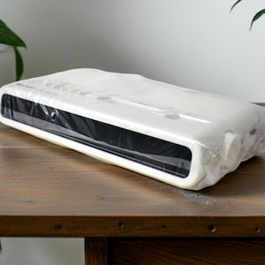 May include: A rectangular electronic device with a white casing and a black front panel, encased in clear protective plastic film. The unit features a series of circular buttons along the top edge and a smooth, rounded design profile.