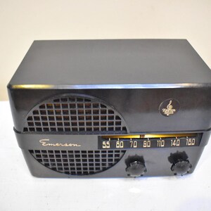 Puede incluir: Una radio Emerson negra vintage con una rejilla de altavoz curvada y un dial de sintonizaci&oacute;n. La radio tiene forma rectangular con bordes redondeados y un peque&ntilde;o emblema en la esquina superior derecha. El dial muestra frecuencias de radio.