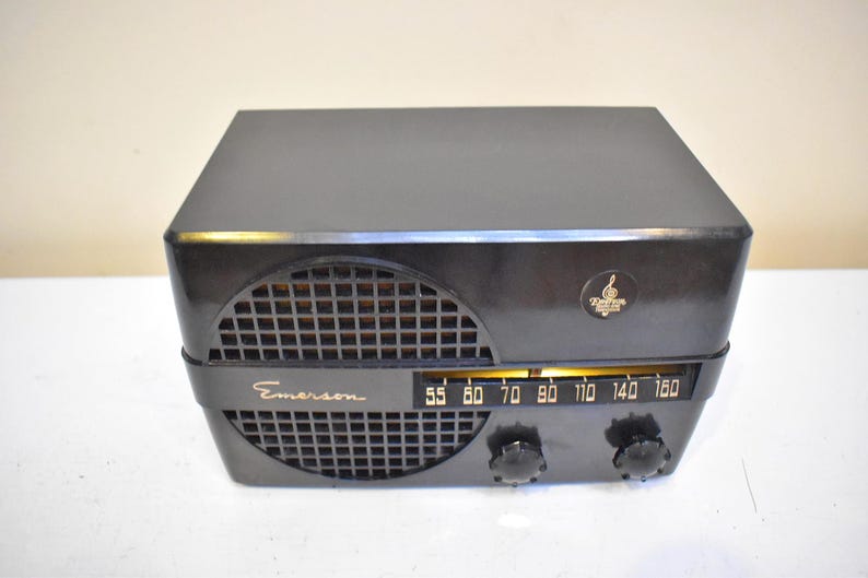 Puede incluir: Una radio Emerson negra vintage con una rejilla de altavoz curvada y un dial de sintonizaci&oacute;n. La radio tiene forma rectangular con bordes redondeados y un peque&ntilde;o emblema en la esquina superior derecha. El dial muestra frecuencias de radio.