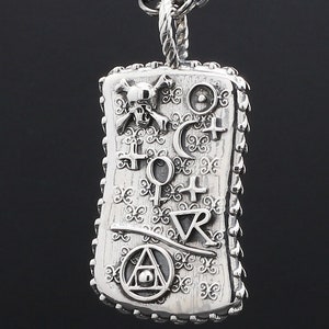 May include: A silver pendant necklace with a rectangular charm featuring a skull and crossbones, a crescent moon, a circle with a dot in the center, a triangle with a circle and dot in the center, and other symbols. The charm has a textured surface and a decorative border.