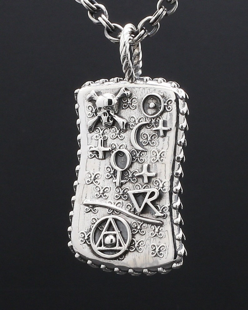 May include: A silver pendant necklace with a rectangular charm featuring a skull and crossbones, a crescent moon, a circle with a dot in the center, a triangle with a circle and dot in the center, and other symbols. The charm has a textured surface and a decorative border.
