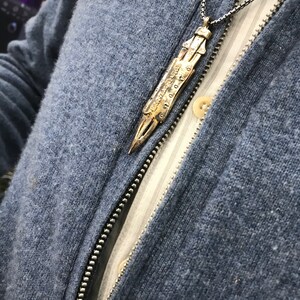 Puede incluir: Un collar con un colgante dorado de dise&ntilde;o detallado e intrincado. El colgante tiene forma de una hoja larga y delgada con una punta puntiaguda. El collar se lleva sobre un su&eacute;ter azul.