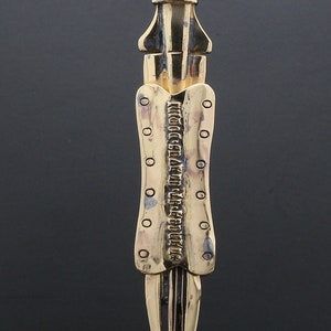 Puede incluir: Un colgante dorado con forma de daga con una cadena de plata. El colgante tiene un dise&ntilde;o detallado y una inscripci&oacute;n que dice "Wicca's Garden of Life Eternal".