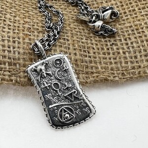 May include: A silver necklace with a pendant featuring an intricate design of symbols and a skull. The pendant is rectangular and has a textured surface. The necklace is on a burlap background.