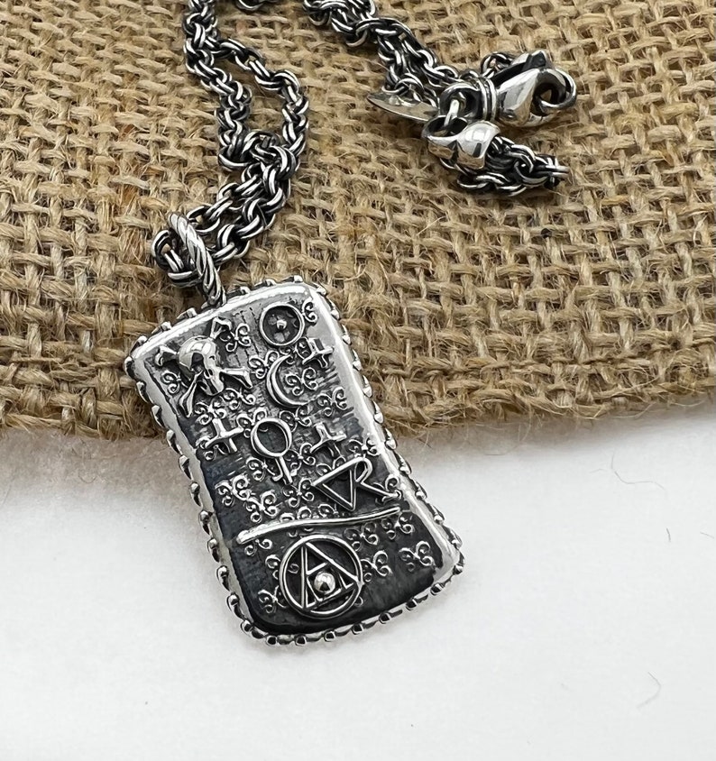 May include: A silver necklace with a pendant featuring an intricate design of symbols and a skull. The pendant is rectangular and has a textured surface. The necklace is on a burlap background.