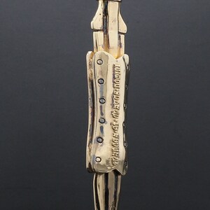 Puede incluir: Collar con colgante dorado con un dise&ntilde;o de cuchillo detallado con una superficie texturizada y una inscripci&oacute;n. La inscripci&oacute;n dice "THE FUTURE IS NOW, SO DO IT NOW".