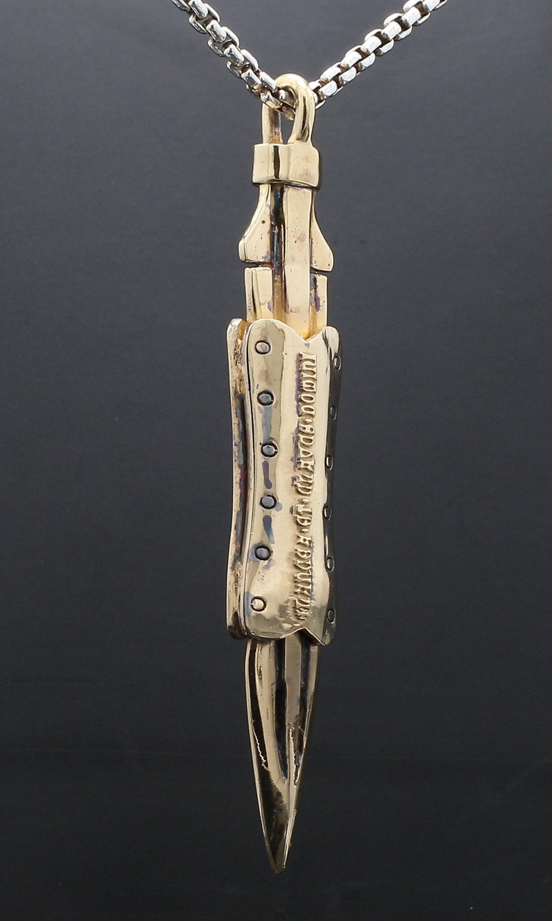 Puede incluir: Collar con colgante dorado con un dise&ntilde;o de cuchillo detallado con una superficie texturizada y una inscripci&oacute;n. La inscripci&oacute;n dice "THE FUTURE IS NOW, SO DO IT NOW".