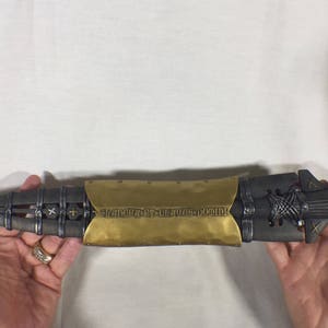 Puede incluir: Una lanza decorativa negra y dorada con un dise&ntilde;o detallado y la inscripci&oacute;n "Strike First, Strike Hard, No Mercy" en la parte dorada.