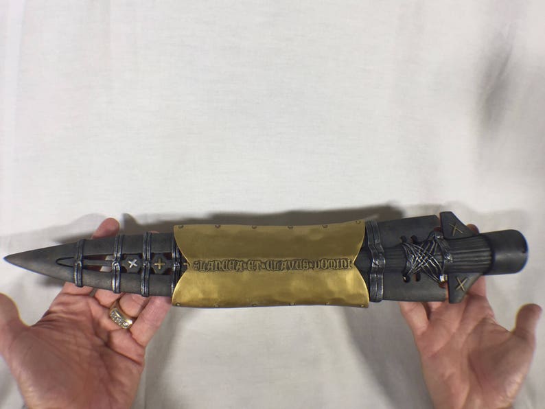 Puede incluir: Una lanza decorativa negra y dorada con un dise&ntilde;o detallado y la inscripci&oacute;n "Strike First, Strike Hard, No Mercy" en la parte dorada.