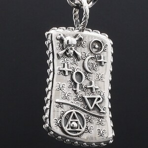 May include: Silver pendant necklace with a rectangular charm featuring a skull and crossbones, a compass, and other symbols. The charm has a detailed, ornate design.