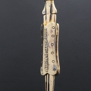 Puede incluir: Un collar con un colgante de color dorado en forma de daga detallada. El colgante tiene la inscripci&oacute;n "TURNING LEAF CO. LTD. LONDON" en el lateral.