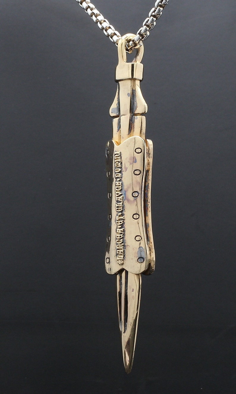 Puede incluir: Un collar con un colgante de color dorado en forma de daga detallada. El colgante tiene la inscripci&oacute;n "TURNING LEAF CO. LTD. LONDON" en el lateral.