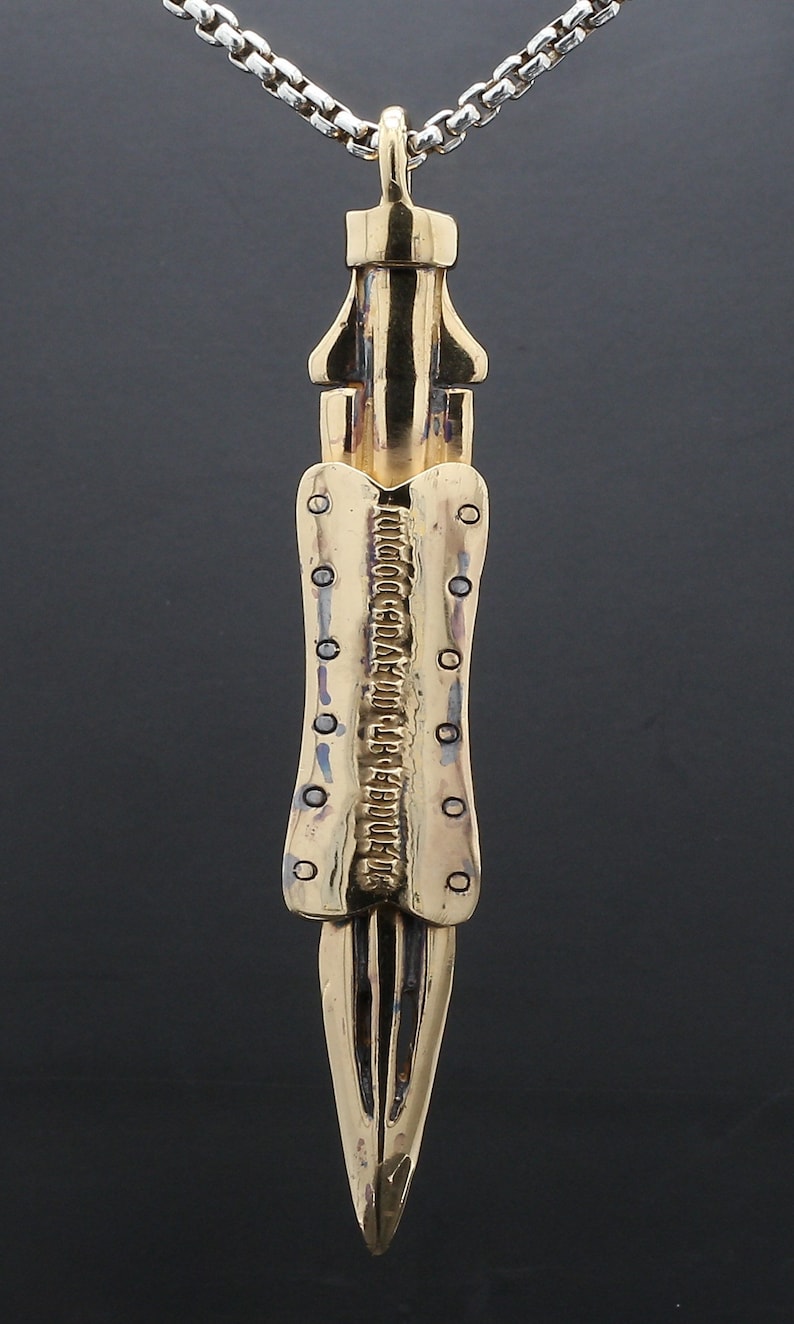 Puede incluir: Un collar con un colgante en forma de espada, de color dorado, con una superficie texturizada. La cadena es de plata.