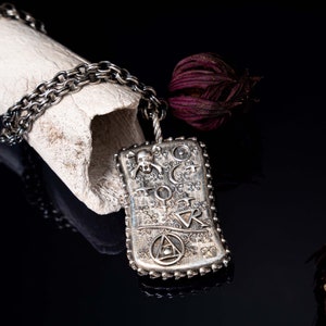 May include: A silver pendant necklace with a rectangular charm featuring a skull, a crescent moon, a triangle, and other symbols. The charm is attached to a silver chain.