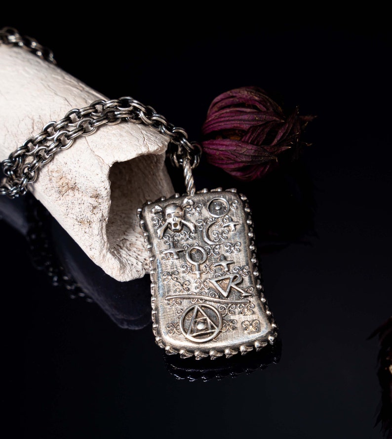 May include: A silver pendant necklace with a rectangular charm featuring a skull, a crescent moon, a triangle, and other symbols. The charm is attached to a silver chain.
