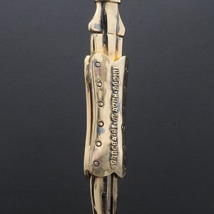 Puede incluir: Un collar con un colgante en forma de daga detallada, de color dorado, con una superficie texturizada y una inscripci&oacute;n. El colgante est&aacute; unido a una cadena plateada.