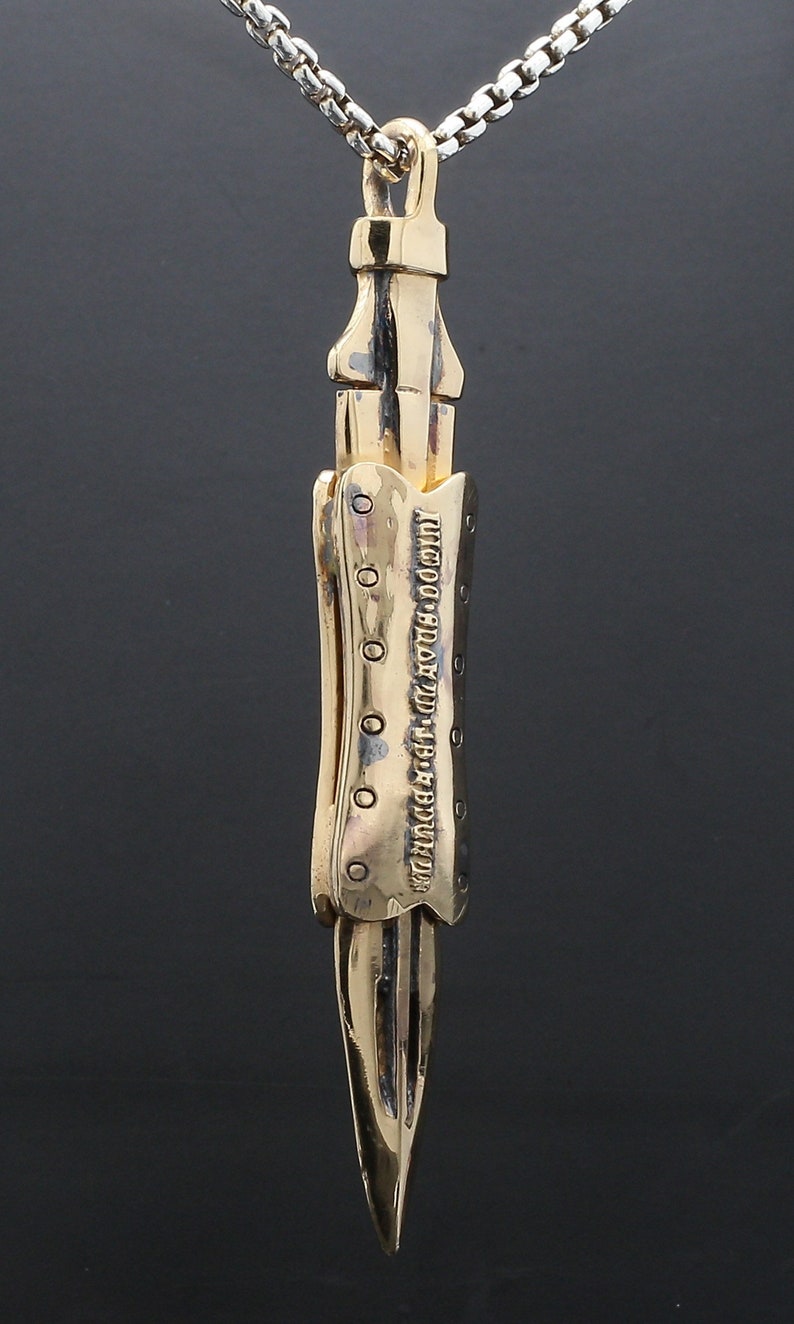 Puede incluir: Un collar con un colgante en forma de daga detallada, de color dorado, con una superficie texturizada y una inscripci&oacute;n. El colgante est&aacute; unido a una cadena plateada.