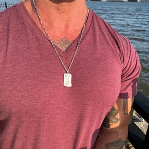 May include: A silver chain necklace with a small rectangular pendant. The pendant has a textured design and is worn by a person with a burgundy t-shirt.