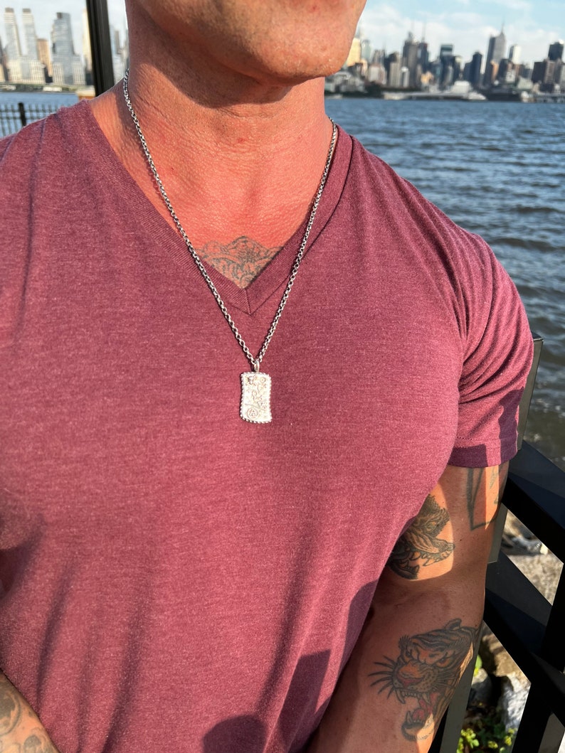 May include: A silver chain necklace with a small rectangular pendant. The pendant has a textured design and is worn by a person with a burgundy t-shirt.