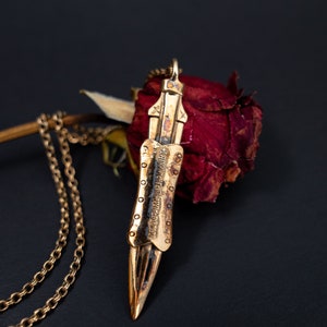 Puede incluir: Un collar de oro con un colgante en forma de espada. El colgante tiene un dise&ntilde;o detallado y una inscripci&oacute;n. El collar est&aacute; sobre una rosa roja seca.