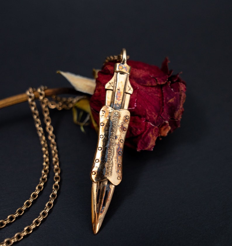 Puede incluir: Un collar de oro con un colgante en forma de espada. El colgante tiene un dise&ntilde;o detallado y una inscripci&oacute;n. El collar est&aacute; sobre una rosa roja seca.