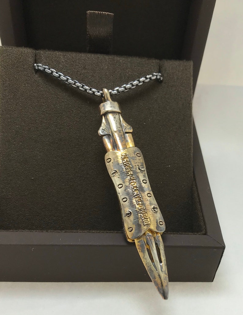 Puede incluir: Un collar de cadena de plata con un colgante de plata y oro con forma de cuchillo. El colgante tiene una inscripci&oacute;n detallada que dice "LEGENDA ARTIS AVE DOCTOREM".