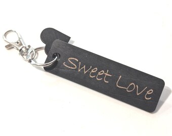 May include: A black wooden keychain with the words "Sweet Love" engraved in a cursive font. The keychain has a silver-colored metal clasp and a small heart-shaped tag attached to the wooden rectangle.