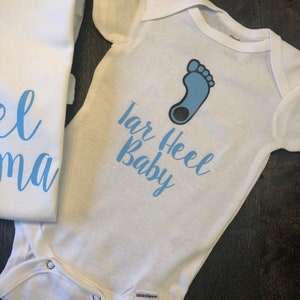 May include: White baby onesie with a blue footprint and the text "Tar Heel Baby" in blue lettering.