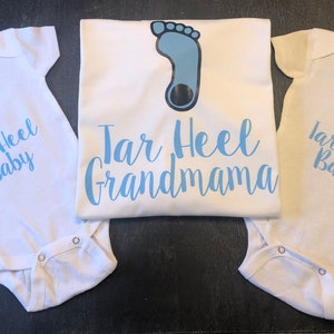 May include: Three white baby onesies with a blue footprint and the text "Tar Heel Baby" on two of them. The third onesie has the text "Tar Heel Grandmama".