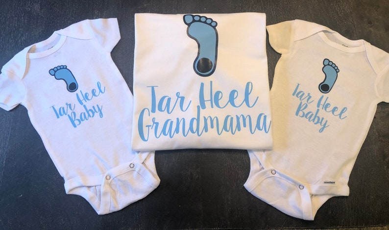 May include: Three white baby onesies with a blue footprint and the text "Tar Heel Baby" on two of them. The third onesie has the text "Tar Heel Grandmama".