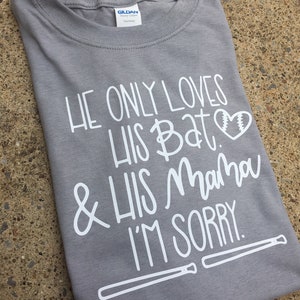 May include: Grey t-shirt with white text that reads "He only loves his bat & his mama I'm sorry." A white baseball bat graphic is at the bottom of the shirt.