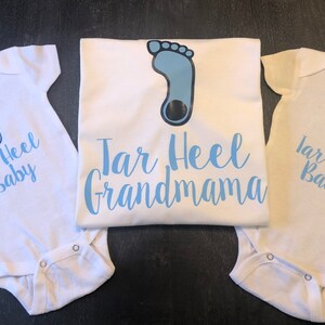 May include: Three white shirts with blue text and a blue footprint design. The shirts say "Tar Heel Baby" and "Tar Heel Grandmama".