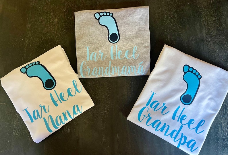 May include: Three white t-shirts with a blue footprint graphic and the text "Tar Heel" followed by "Grandma", "Nana", and "Grandpa".