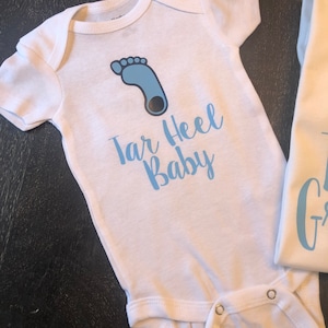 May include: White baby onesie with a blue footprint and the text "Tar Heel Baby" in blue.