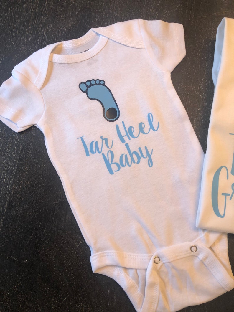 May include: White baby onesie with a blue footprint and the text "Tar Heel Baby" in blue.