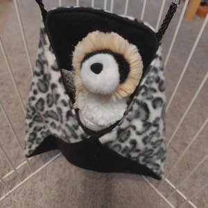 May include: A hanging pet hammock featuring a black and white leopard print fleece exterior with a solid black bottom section. The hammock is suspended by black braided cords and metal clips, containing a small plush toy with a tan and white face.