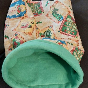 May include: A fabric pouch featuring a beige background with a repeating Christmas-themed pattern of snowmen, decorated pine trees, and holly leaves. The interior of the pouch is lined with a solid, soft-textured light green fleece fabric.