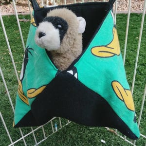 May include: A hanging fabric pouch featuring a green background with a yellow duck pattern and black accents. A plush ferret toy with tan fur and a black mask around its eyes is positioned inside the pouch, which is suspended by two black straps.