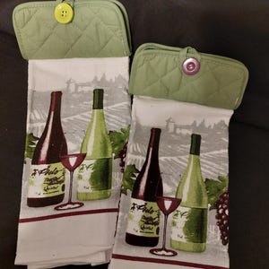 May include: Two white kitchen towels with a green quilted pot holder on top of each. The towels feature a wine theme with a bottle of red wine, a bottle of white wine, and a glass of red wine. The text on the bottles reads "F. Piccolo, Premier Qualite".
