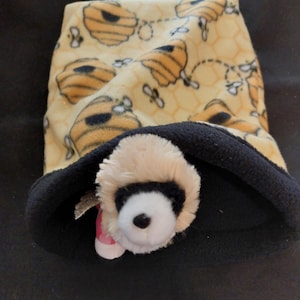 May include: A soft, yellow fleece pouch featuring a repeating pattern of brown beehives and small bees. The interior is lined with solid black fleece. A small, plush animal head with cream-colored fur and a white face is peeking out from the opening.