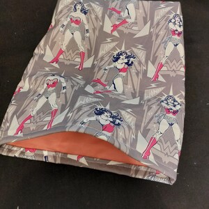 May include: A gray fabric item featuring a repeating pattern of Wonder Woman illustrations in her classic costume. The interior lining is a solid orange color. The design includes lightning bolt shapes and the word "Wonder Woman".