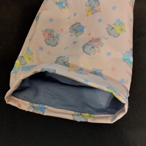 May include: A light pink baby blanket with a pattern of cartoon elephants, stars, and hearts. The blanket has a blue interior and is folded over, revealing the inside. The elephants are in various colors, including blue, yellow, and red.