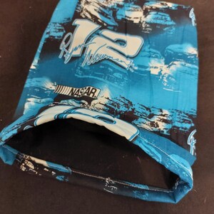 May include: A blue and black fabric item featuring the number 12 and the name "Ryan Newman" in white. The word "NASCAR" is also visible. The item has a pocket and is likely a bag or pouch.