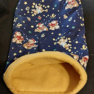 May include: A fabric tube with a dark blue exterior featuring a repeating pattern of teddy bears in Santa hats, angels with trumpets, yellow stars, and rocking horses. The interior is lined with a solid mustard yellow fleece fabric.