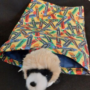 May include: A plush ferret toy peeks out of a fabric pouch. The pouch is yellow and covered in a crayon pattern with the word "Crayola" printed on the crayons. The ferret has a tan and black fur pattern.