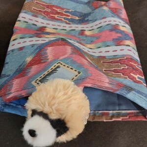 May include: A folded fabric item with a Southwestern pattern in shades of blue, red, and brown. A small, plush animal with a black and white face peeks out from the fabric. The item appears to be a decorative accessory.