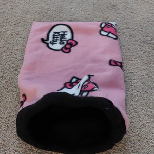 May include: Pink fleece pet bed with a black interior. The bed features a repeating pattern of Hello Kitty characters and speech bubbles. The speech bubbles say "Hello Kitty" with a pink bow. The bed is designed for small pets.