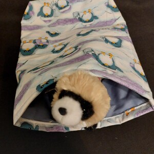 May include: A fabric pouch with a penguin pattern in shades of blue, purple, and white. A stuffed animal with a tan and black fur is nestled inside the pouch. The pouch is open, revealing a blue interior.